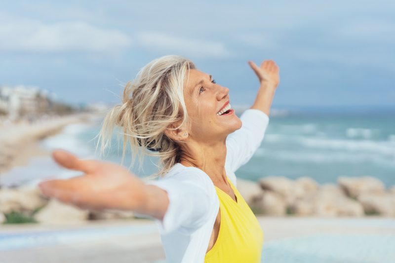 Joyful woman enjoying the freedom of the beach standing with open arms and a happy smile looking up towards the sky